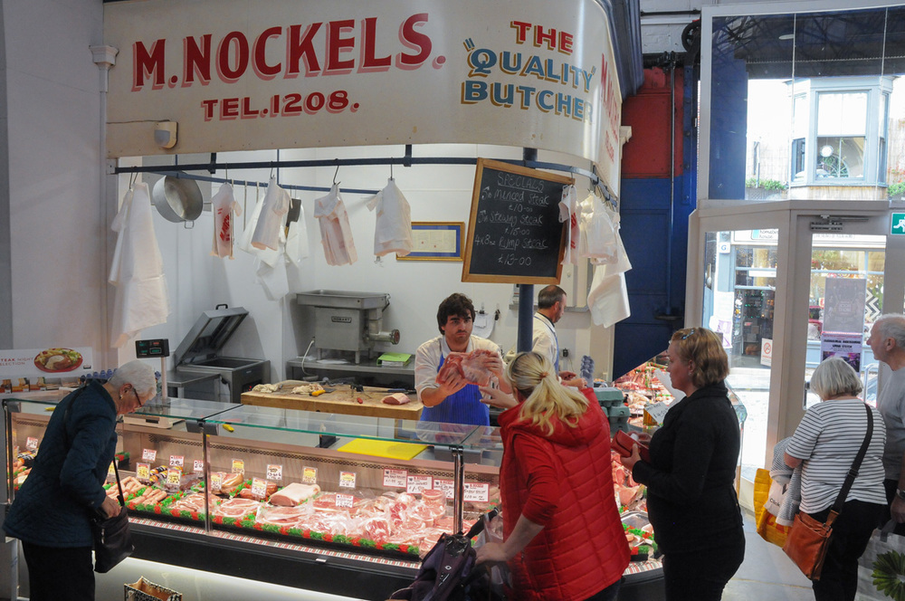 The M. Nockels counter inside Scarborough Market Hall — the painted fascia sign above a full glass display counter, aprons hanging from the rail and customers being served